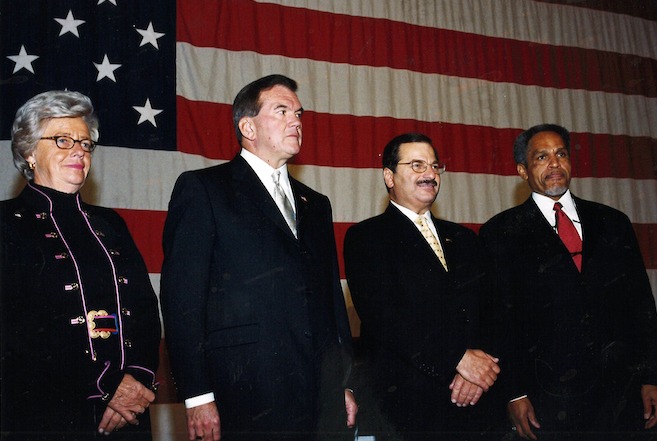 Left to right: Judee von Seldeneck, PA Governor Tom Ridge, Aqua CEO Nick DeBenedictis and Philadelphia Mayor John Street. A very tanned white woman stands alongside three men in suits: One is white, one is white with a mustache, and one is Black with a beard — all in front of a large American flag.