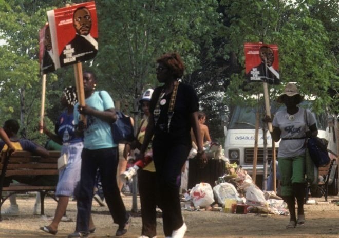 African Americans walk in a park in Washington, D.C. with signs depicting Martin Luther King, Jr. in 1983. Behind them, trash overflows from a trash can.