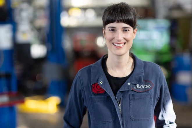 Washington State Rep. Marie Gluesenkamp Perez, a white woman with dark brown bangs, wears a zip-up mechanic suit with her name embroidered on the chest and a rose embroidered on the other side.