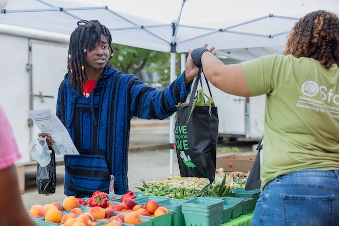 At an outdoor farmstand in Nicetown-Tioga, beneath a white ten, a Black person wearing beaded locs and a striped woven hoodie receives a tote bag of produce from a Pennsylvania Horticultural Society Worker in a green t-shirt and jeans. Between them is a table with paper basketbs of peaches, red bell peppers and more produce.