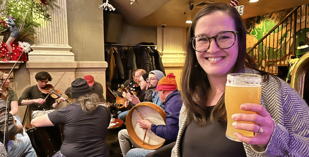 Rosaleen McGill, a white woman with brown straight beyond-shoulder-length hair, large tortoise shell glasses, a tweed sweater and black top, holds a pint of beer in The Plough & The Stars, an Irish pub in Old City, Philadelphia. Behind her are a group of white musicians playing.