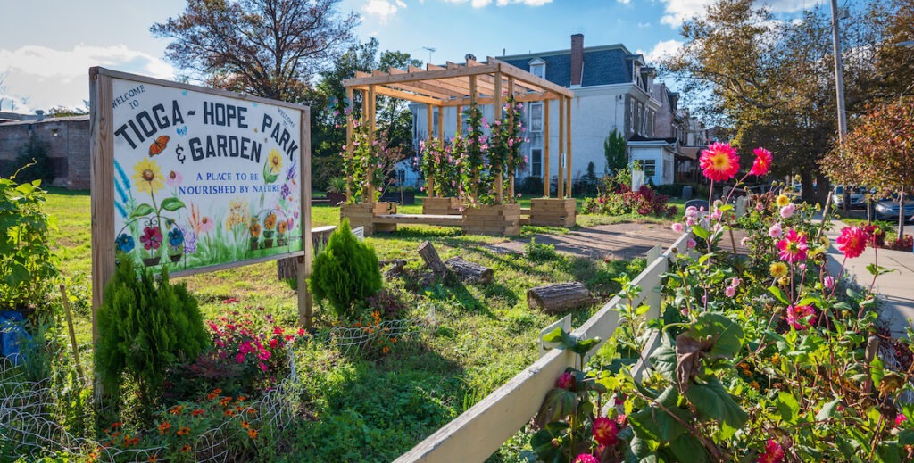 A sign painted with flowers stands behind a bed of blooming flowers and in front of a green lawn. The sign reads "WELCOME TO TIOGA-HOPE PARK & GARDEN A PLACE TO BE NOURISHED BY NATURE." Row homes stand in the far ground beyond the park.