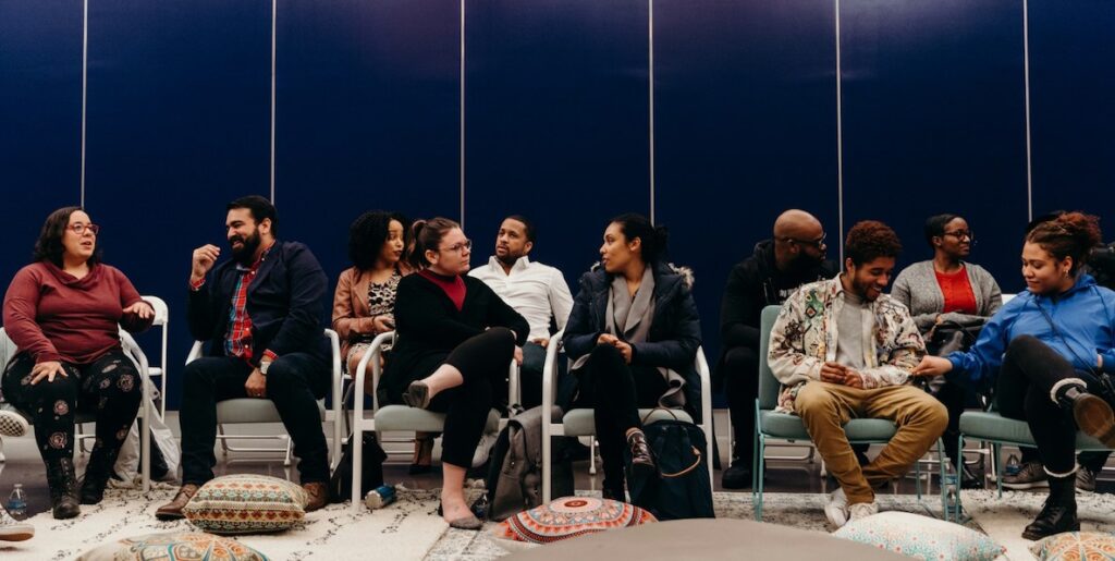 A diverse group of adults sit in two lines of chairs that face front. At their feet are pillows.