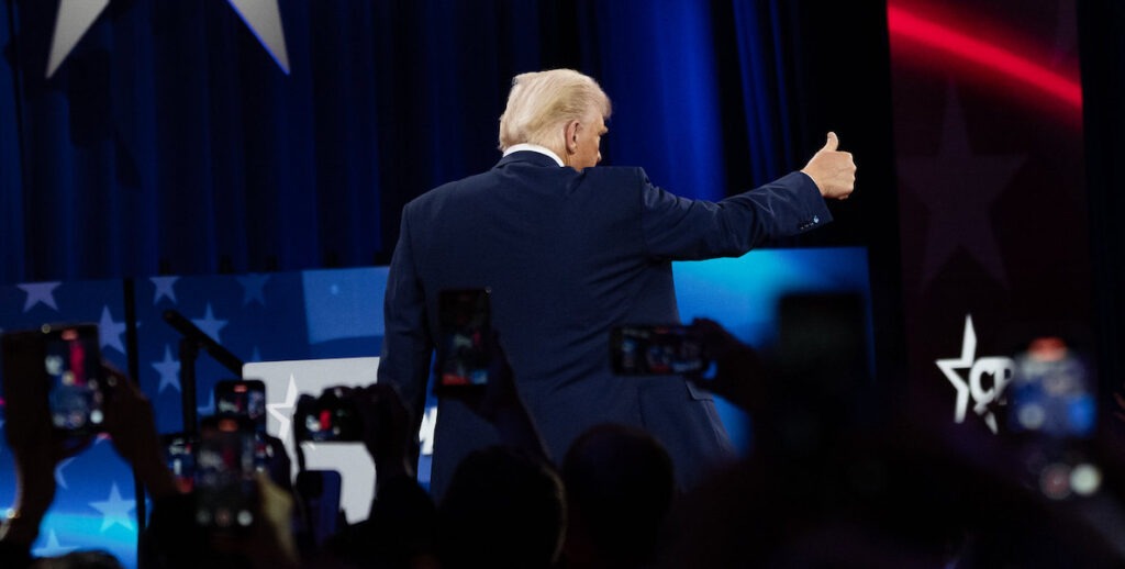 The back of President Donald J. Trump wearing a navy suit gives the thumbs up at CPAC 2025.