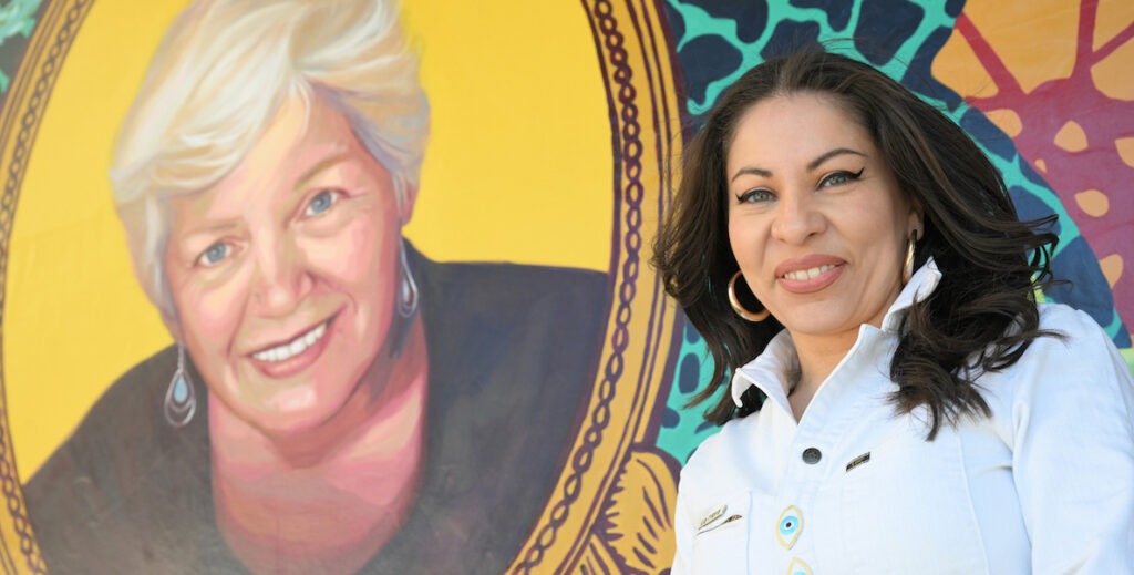 Artist Michelle Angela Ortiz stands beside a mural of a white woman with short blonde hair entitled "Our Market Ancestor: Betty Ann Mongelluzo."