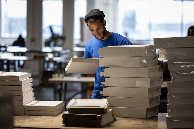 Baker Industries participant Angel, a man with a short black bear, black beanie and blue t-shirt, stacks bocks on a table as he works on an assembly line.
