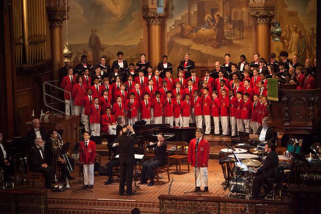 Three rows of boys wearing red blazers and white pants, white shirts and black ties and another row of adult singers behind them in black jackets, white shirts and red bowties stand on a stage and sing. A small band plays in front of them. Two boys stand at the front of the stage singing into microphones.