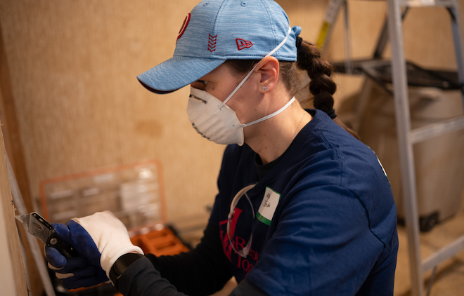 A white woman in a blue cap with a long braid wearing an N-95 mask and blue shirt and white gloves scrapes at a wall while repairing a home.