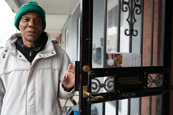 Rudolph Edge, a Black man in a green beanie and khaki jacket, stands on a porch, holding a metal-frame storm door open.