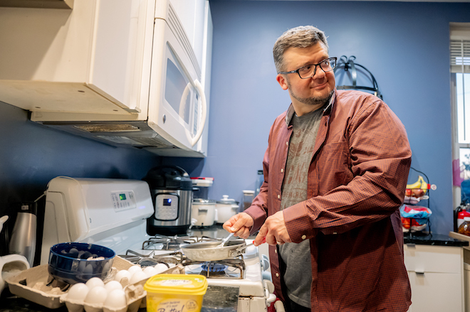Joe Gidjunis, a White man with grey hair and a short beard wearing an open-button down shirt with a t-shirt under it, stands at a stovetop to cook eggs in a pan.