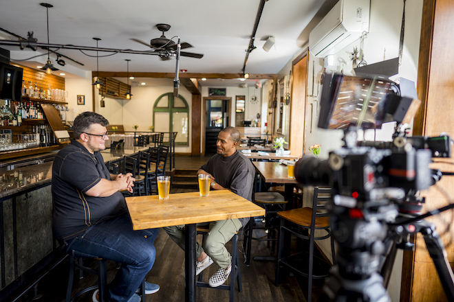 Joe Gidjunis, a White man with grey hair and a short beard wearing a dark shirt, jeans and sneakers, sits across a tall bar table (and two pints of beer) from Jeremy Givens, a Black man with short hair wearing a dark shirt, cargo pants and checkered vans. A camera in the foreground records the conversation.