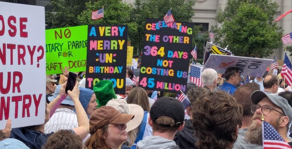 A crowd of people's heads, shoulders, and some arms are is gathered outside in front of Philadelphia's Suburban Station, many are holding protest signs. The two central to the image are black posterboard with rainbow lettering, one which reads "A very merry un-birthday to you" and the other reads "Celebrating the other 364 days not costing us 45 million"