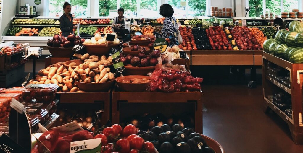 Women shop near a window inside the produce section of a grocery store.