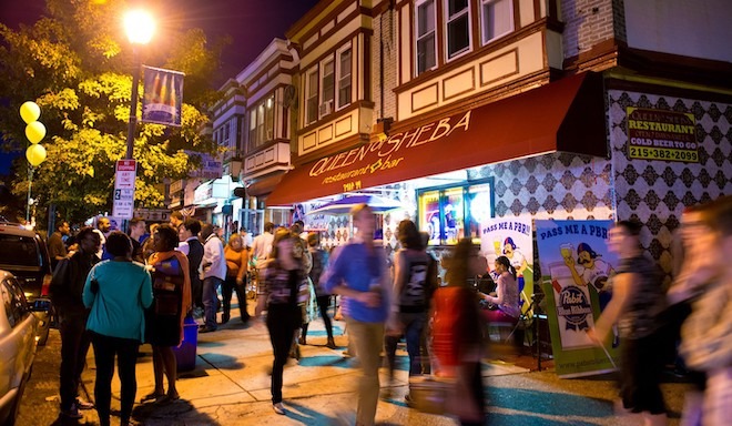 People walk along a city sidewalk during an evening Baltimore Avenue Dollar Stroll in West Philadelphia.