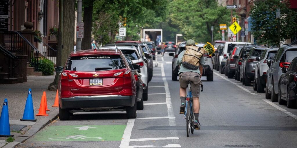 A person rides a bike in the middle of a Philadelphia street, toting a back with flowers in it on his back. To his left, cars are parked in a designated bike lane.