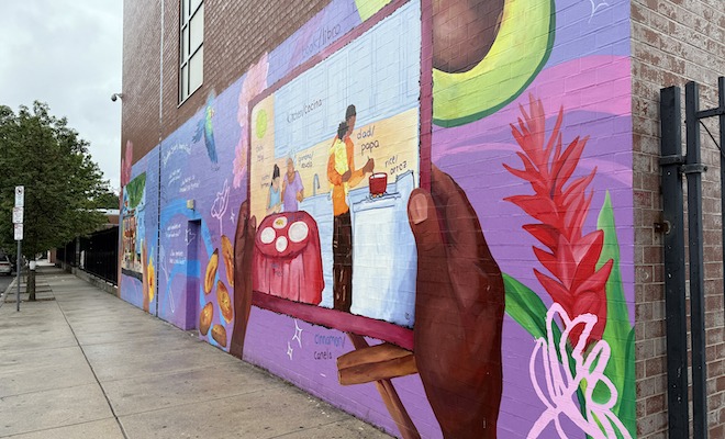 A bright mural outside Julia de Burgos Elementary School depicts a brown hair holding a picture of a family eating and cooking.
