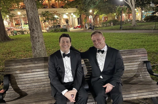 Tim Judge and Kevin Hart, two white men wearing tuxedos, sit on a wooden bench in Rittenhouse Square.