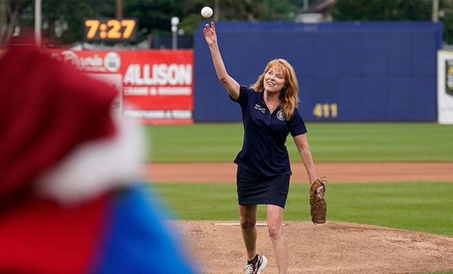 Pennsylvania State Treasurer Stacy Garrity, a petite white woman with fluffy strawberry blonde bangs and feathered long hair in a short blue shirt and navy blouse, throws out the first pitch at a minor league baseball game in Williamsport, PA.