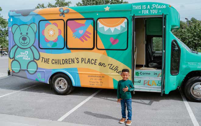 A small African American child stands in front of a colorful mental healthcare bus called The Children's Place On Wheels.