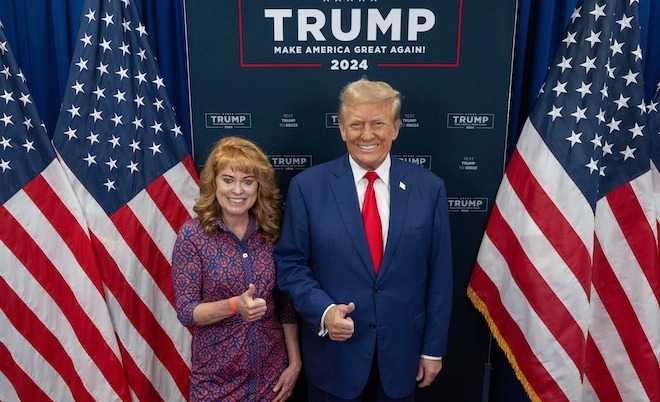 Pennsylvania State Treasurer Stacy Garrity, a petite white woman with fluffy strawberry blonde bangs and feathered long hair in a blue and red button-down dress, stands in front of U.S. flags and a TRUMP Make America Great Again 2024 sign, alongside Donald Trump, a taller man with frosted blonde hair in an oversize navy suit, white shirt and red tie. Both are smiling and putting their right thumbs up.