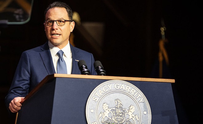 Governor Josh Shapiro, a white man with dark brown hair, glasses, a navy jacket, white shirt, grey-blue tie, stands behind a podium bearing the seal of Pennsylvania and the words "STACY GARRITY" at Garrity's 2024 inauguration.