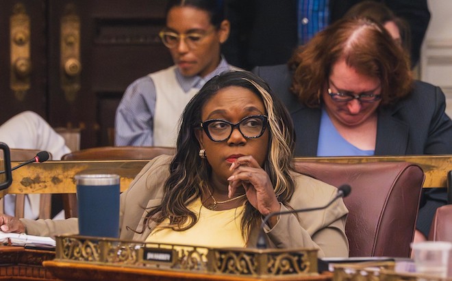 Kendra Brooks, a Black woman with long hair, a blonde streak, glasses, wearing a khaki jacket with a yellow shirt, sits in a Committee of the Whole hearing about the 2026 budget in City Hall. 