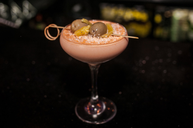 A milky "dirty Caesar" martini, made of Caesar dressing and alcohol, comes in a martini glass, with grated parmesan and speared green olives on top.