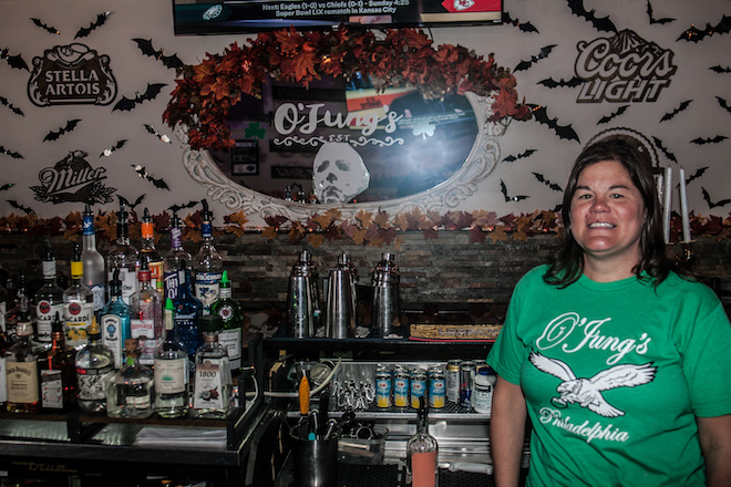 Holly Otto, a White woman with shoulder length brown hair wears a green t-shirt that says "O'Jung's Philadelphia" and has the Eagles logo while standing behind the bar.