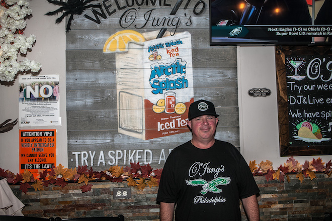 Mike Otto Sr., a white man in a black t-shirt that says "O'Jung's Philadelphia" and has the Eagles logo, and a black O'Jung's baseball cap stands in front of a mural depicted an Artic Splash Iced Tea.
