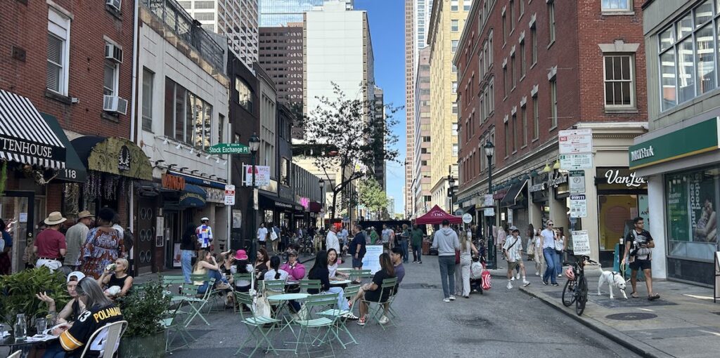 People sit outside around cafe tables along West Walnut Street in Philadelphia. Skyscrapers show in the background.