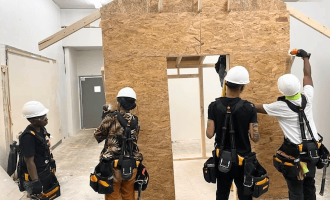 For people in white hard hats and work belts with suspenders work on a particle-board wall.