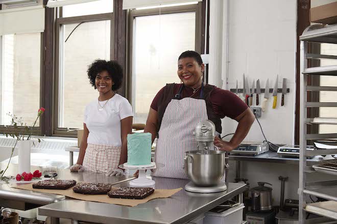 Mercedes Brooks (left) and Rhonda Saltzman, two Black women, one in an apron, stand among baked goods, a mixer, knives and other kitchen supplies in Second Daughter bakery.