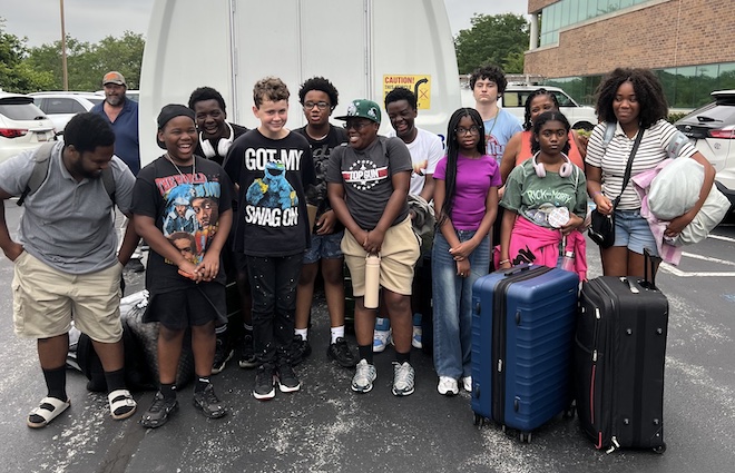 Mostly Black teenagers in actual wear stand, some with luggage in front of the back of a white van. They are participants in the YMWIC, going on a college campus tour.