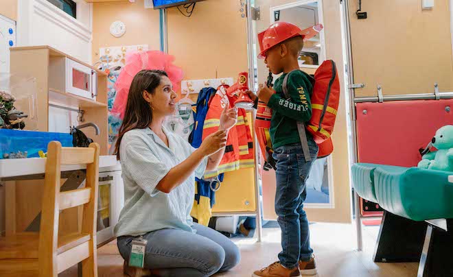 A small African American child in jeans, a long-sleeve shirt and toy firefighter's helmet and jacket stands facing a woman with long hair, a loose blouse and jeans who is a mental healthcare worker. They are inside a free mental health service bus for children called The Children's Place On Wheels.