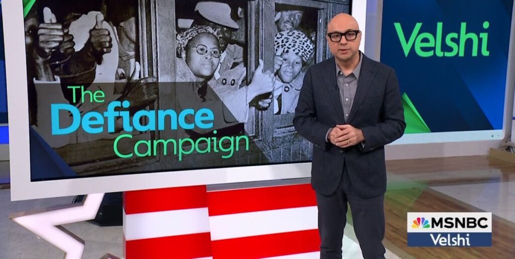 A bald man in glasses and a navy suit stands before a monitor showing detained South Africans crowded inside a vehicle in the 1950s and a graphic that reads "The Defiance Campaign"