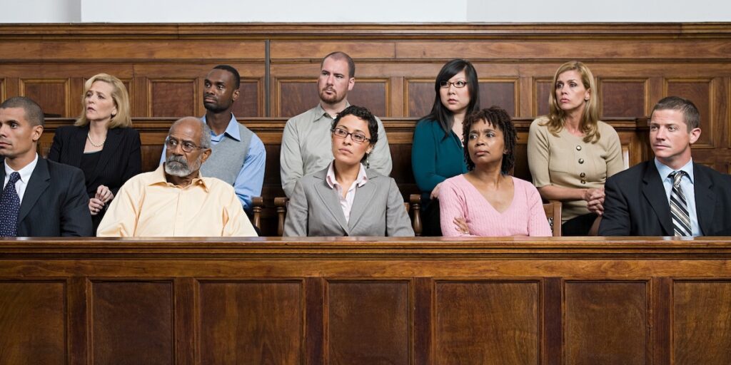 A jury of diverse adults, all dressed in business attire, sits in a jury box behind a wooden panel, all looking seriously in the same direction.